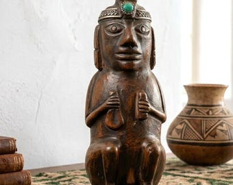 Puede incluir: Una estatua de madera tallada marrón de una figura sentada, sosteniendo objetos en sus manos. La estatua tiene un tocado con una piedra turquesa y está junto a un jarrón decorativo. La estatua está sobre una mesa con una alfombra estampada.