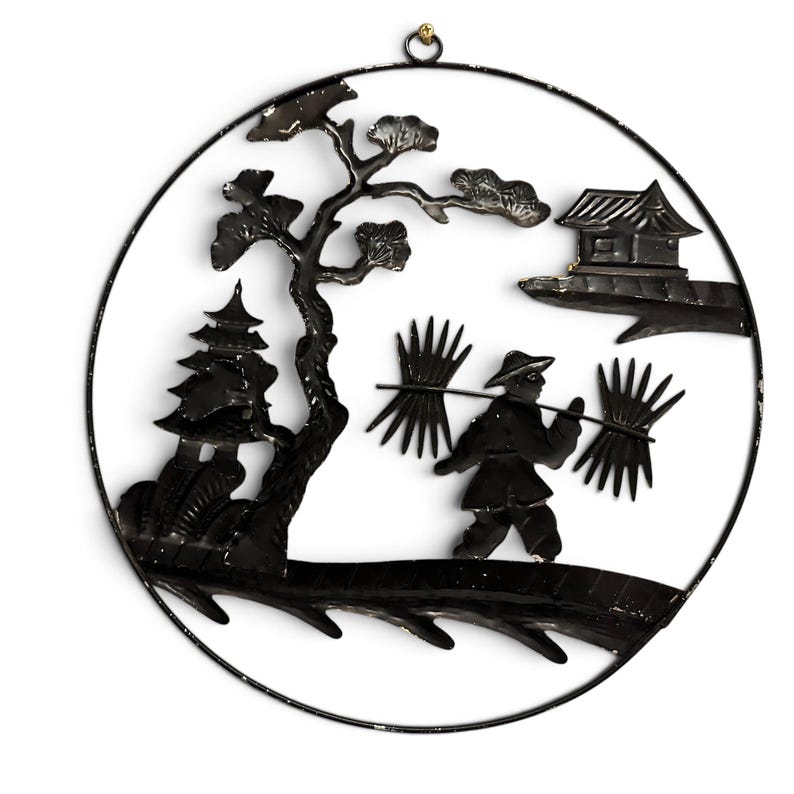May include: A circular black metal wall hanging featuring a silhouette design of a person carrying bundles on a pole across their shoulders, a tree, a small building, and a pagoda structure, all enclosed within a thin black metal circular frame.