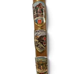Puede incluir: Un bast&oacute;n de madera con tres escudos met&aacute;licos de recuerdo. El escudo superior representa una escena de monta&ntilde;a con el texto Flexenstrasse. El escudo central muestra una carretera sinuosa con el texto Arlberg-Passtrasse. El escudo inferior muestra un arco con el texto Innsbruck.
