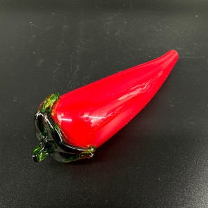 May include: A glass sculpture shaped like a chili pepper with a vibrant red body and a dark green stem. The surface of the glass is smooth and reflective, showcasing a tapered, elongated form that curves slightly toward the pointed end.