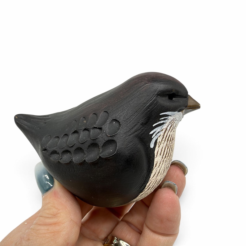 May include: A small, rounded bird figurine crafted from dark brown material. The surface features carved, scale-like wing details and painted white, textured markings on the chest area. The bird has a pointed, light brown beak.