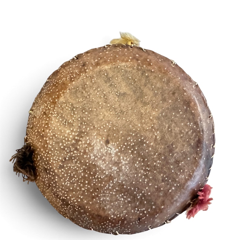 May include: A circular, brown, textured drum head featuring a speckled white pattern across its surface. The edges are secured with visible stitching, and small tufts of yellow, brown, and pink fibres are attached to the perimeter of the frame.
