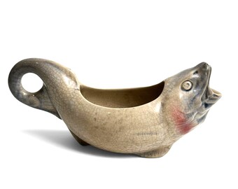 Może przedstawiać: Ceramiczne naczynie w kształcie ryby, pełniące funkcję sosjerki lub dzbanka, z beżowo-szarym szkliwem typu krakle. Naczynie posiada szeroko otwarty pysk służący za wylew, uchwyt w kształcie zakrzywionego ogona oraz różowo-czerwony akcent przy skrzelach.