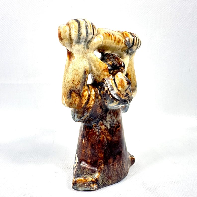 May include: A ceramic figurine with a brown and tan glaze. The figurine depicts a figure holding a smaller figure. The base is a dark brown color. The figurine has a glossy finish and is set against a white background.