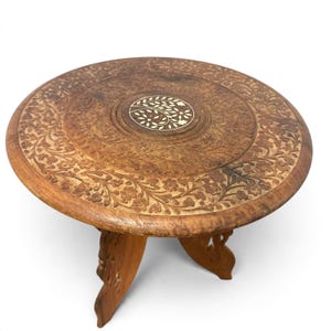 May include: A round wooden table with a dark brown finish, featuring intricate floral carvings across the surface and a central circular inlay with a white leaf pattern. The table is supported by three carved, folding wooden legs.