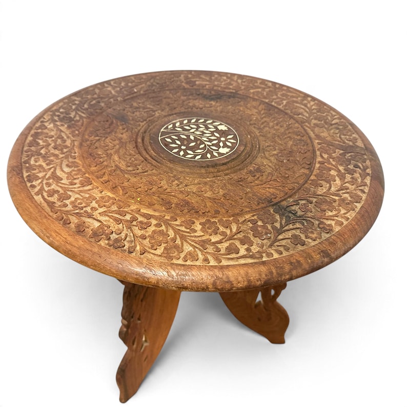 May include: A round wooden table with a dark brown finish, featuring intricate floral carvings across the surface and a central circular inlay with a white leaf pattern. The table is supported by three carved, folding wooden legs.