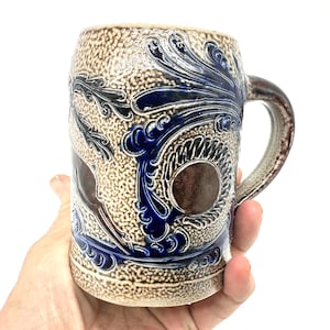 Puede incluir: Una jarra de cerveza de gres esmaltado con sal, con una superficie beige texturizada y patrones en relieve de color azul cobalto, que incluyen volutas y motivos circulares. El recipiente cil&iacute;ndrico tiene un asa resistente y un acento circular liso.