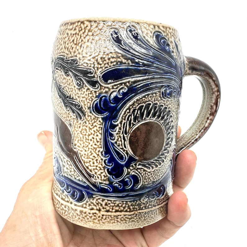 Puede incluir: Una jarra de cerveza de gres esmaltado con sal, con una superficie beige texturizada y patrones en relieve de color azul cobalto, que incluyen volutas y motivos circulares. El recipiente cil&iacute;ndrico tiene un asa resistente y un acento circular liso.