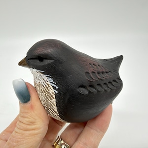 May include: A small, hand-carved wooden bird figurine featuring a dark brown and black finish. The bird has a textured, light-colored chest with carved details, a pointed beak, and carved wing patterns along its side, showcasing a smooth, matte appearance.