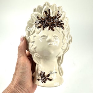 Puede incluir: Jarrón de cerámica color crema con forma de cabeza, con un rostro detallado y una corona floral. El jarrón tiene una superficie texturizada y está adornado con detalles florales marrones. La base tiene un diseño floral marrón y una etiqueta "Rosa Ljung".
