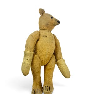 Pode incluir: Um urso de peluche cor de areia com uma superf&iacute;cie de tecido texturizado, apresentando um focinho pontiagudo, olhos e nariz costurados em escuro, e bra&ccedil;os longos que terminam em luvas de croch&eacute; bege claro com detalhes de garras costuradas.