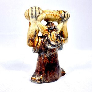 May include: A ceramic sculpture with a figure holding a horizontal object aloft. The sculpture features a color palette of brown, tan, and black. The base is dark brown, transitioning to tan and black details on the upper portion.