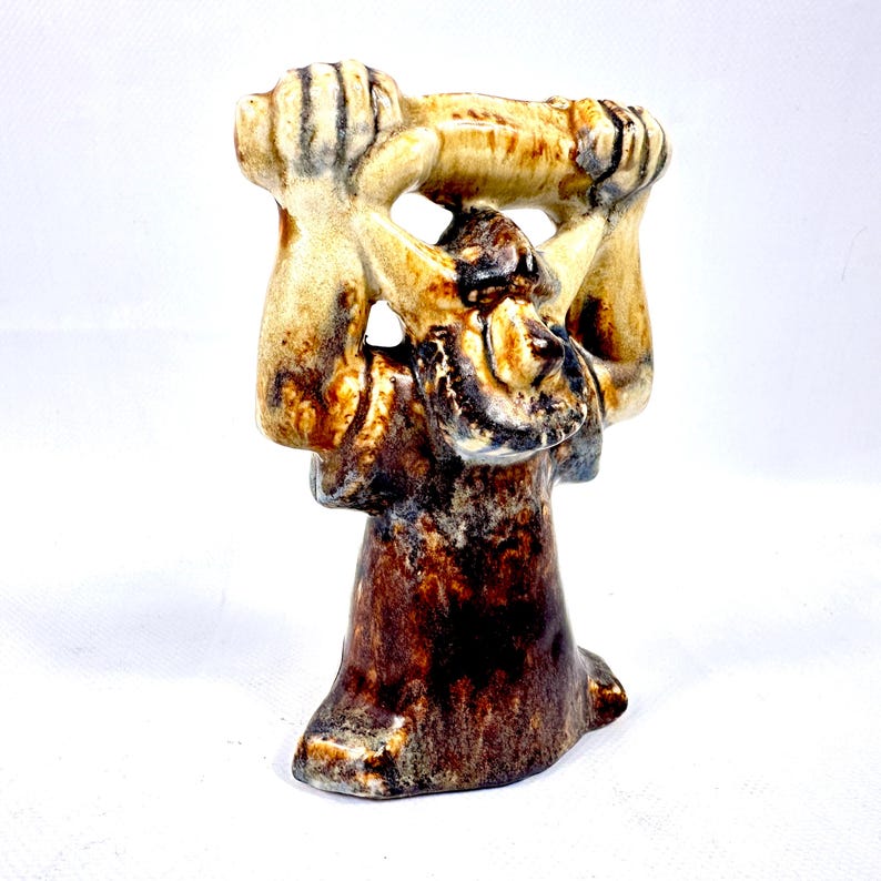 May include: A ceramic sculpture with a figure holding a horizontal object aloft. The sculpture features a color palette of brown, tan, and black. The base is dark brown, transitioning to tan and black details on the upper portion.