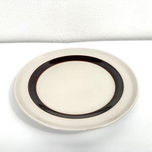 May include: A white ceramic plate with a brown band around the edge. The band has a repeating pattern of small circles.