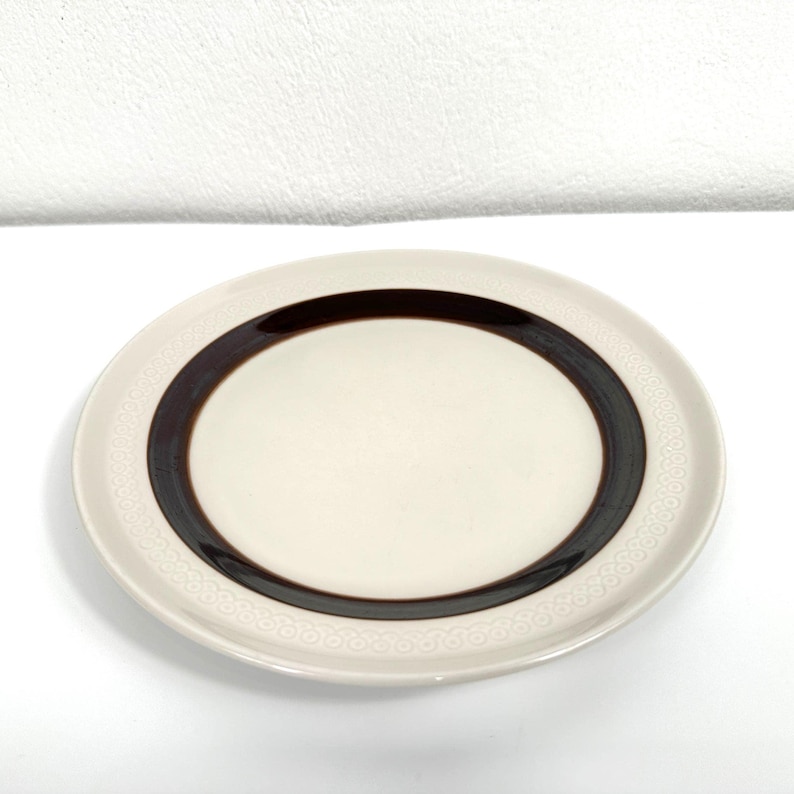 May include: A white ceramic plate with a brown band around the edge. The band has a repeating pattern of small circles.