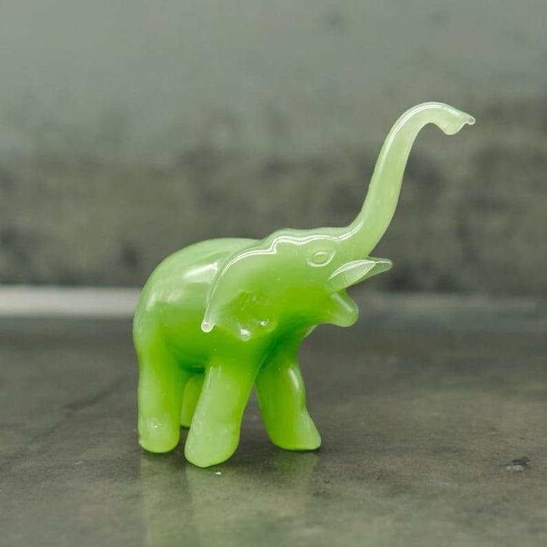 K&ouml;nnte beinhalten: Eine durchscheinende, hellgr&uuml;ne Elefantenfigur mit erhobenem R&uuml;ssel. Die kleine Skulptur hat sichtbare Ohren, Sto&szlig;z&auml;hne und Beine. Der Elefant besteht aus einem glatten, gl&auml;nzenden Material und steht vor einem neutralen Hintergrund.