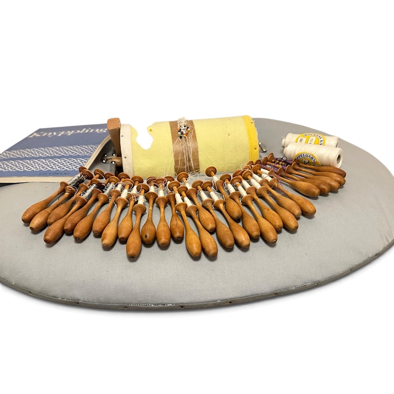 May include: A lace making kit featuring a cylindrical yellow pillow, numerous wooden bobbins with white thread, two spools of white thread, and a blue instructional book with the text Knyppling visible on the cover.