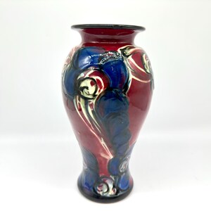 May include: A decorative ceramic vase with a glossy finish. The vase features a maroon base color with blue and white floral designs. The rim and base of the vase are black. The vase is a decorative home decor item.