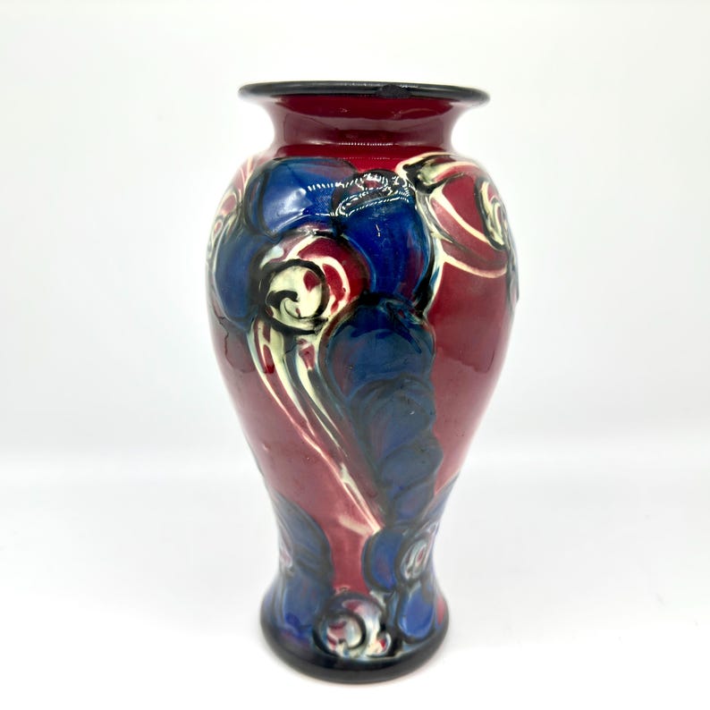 May include: A decorative ceramic vase with a glossy finish. The vase features a maroon base color with blue and white floral designs. The rim and base of the vase are black. The vase is a decorative home decor item.