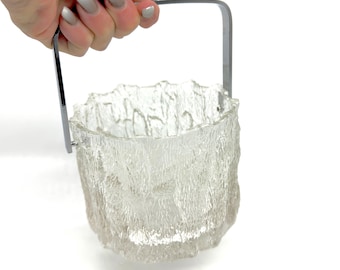 Op de afbeelding: Een heldere, getextureerde glazen ijsemmer met een zilveren metalen handvat. De emmer heeft een ruwe, organische textuur en het handvat is een eenvoudig, gebogen ontwerp. De ijsemmer wordt door een hand omhoog gehouden.