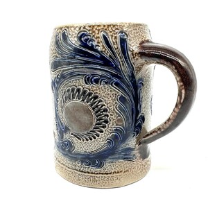 Puede incluir: Una jarra de cerveza cil&iacute;ndrica de cer&aacute;mica con una superficie beige texturizada y un asa de color marr&oacute;n oscuro. La jarra presenta un centro circular prominente rodeado de patrones de hojas en espiral azul oscuro en relieve.