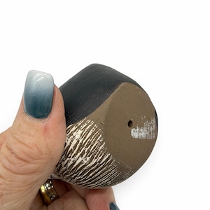 May include: A small, rounded ceramic object with a matte black upper section and a textured, light brown lower section featuring white vertical markings. The base is flat, brown, and includes a single, centered circular drainage hole.