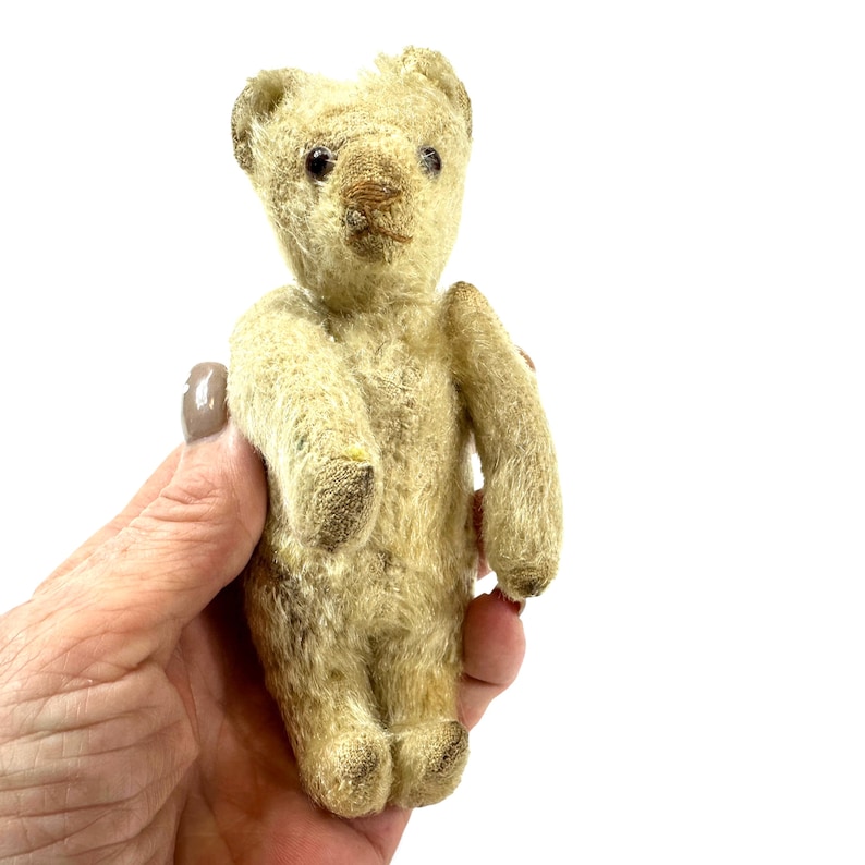 May include: A small, vintage-style teddy bear crafted from light beige, textured mohair-like fabric. The bear features dark, round bead eyes, a stitched brown nose, and small, rounded ears. Its limbs are straight, and the fur appears worn.