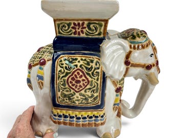 May include: A ceramic elephant garden stool featuring a white body with a glossy finish. The elephant is adorned with a dark blue saddle blanket decorated with intricate green and red floral patterns, gold trim, and a flat rectangular top surface.