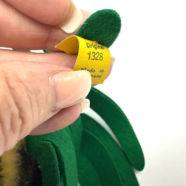 May include: A close-up of a yellow tag with the text "Original 1328 Made in Germany" attached to a green felt object. The tag is held by a person's hand with manicured nails. The object has green felt leaves.