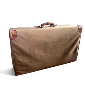 May include: A rectangular brown canvas suitcase featuring a top handle and reinforced leather corner protectors. The exterior fabric shows signs of wear and texture, with a structured frame and a simple, functional design for travel storage.