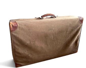 May include: A rectangular brown canvas suitcase featuring a top handle and reinforced leather corner protectors. The exterior fabric shows signs of wear and texture, with a structured frame and a simple, functional design for travel storage.