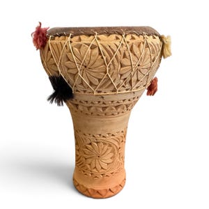 May include: A tan clay goblet drum featuring intricate hand-carved floral patterns across its surface. The drum head is secured with a crisscross cord lacing system and decorated with small, colourful tufts of red, black, and cream-coloured yarn.