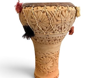 May include: A tan clay goblet drum featuring intricate hand-carved floral patterns across its surface. The drum head is secured with a crisscross cord lacing system and decorated with small, colorful tufts of red, black, and cream-colored yarn.