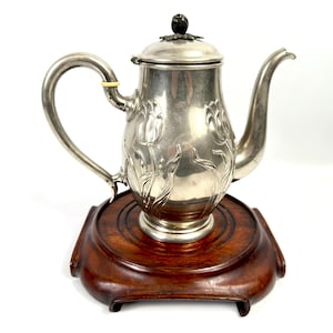 May include: A silver teapot with a tulip floral design, presented on a dark wooden base. The teapot features a curved handle and spout. The lid is topped with a dark, round finial. The wooden base is a dark brown colour.