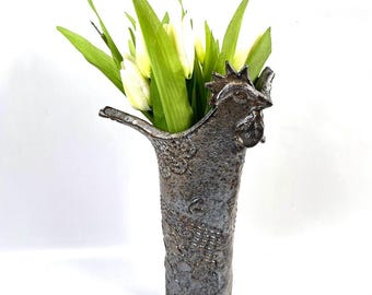 May include: A rustic, textured ceramic vase with a rooster head design. The vase is filled with white tulips and green leaves. The vase has a dark, mottled finish and is suitable for floral arrangements.