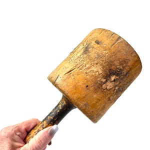 May include: A vintage wooden mallet featuring a large, cylindrical, light brown head with visible surface wear and a dark, weathered handle. The tool displays a natural grain texture and signs of age, with a sturdy construction and a cylindrical striking surface.