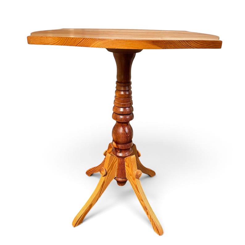 May include: A wooden pedestal table featuring a smooth, light brown oval top supported by a central turned column. The base consists of three curved, splayed legs that provide stability, showcasing a natural wood grain pattern throughout.