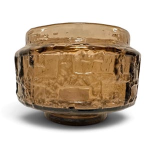 May include: A short, wide, amber-colored glass bowl featuring a textured, block-like relief pattern on its exterior. The glass has a translucent, brownish hue with a smooth, rounded rim and a circular base, creating a sturdy, cylindrical form.