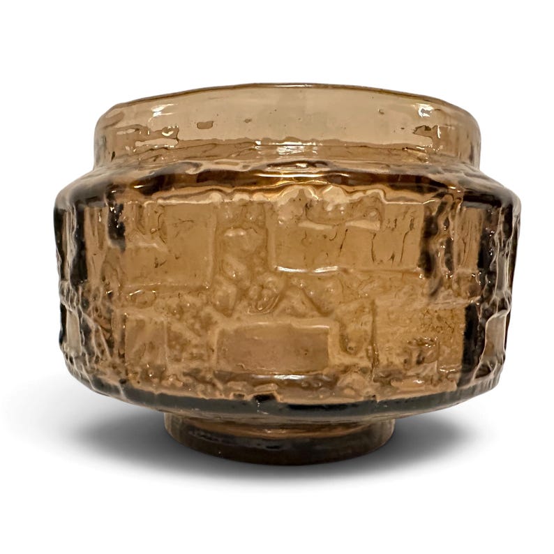 May include: A short, wide, amber-colored glass bowl featuring a textured, block-like relief pattern on its exterior. The glass has a translucent, brownish hue with a smooth, rounded rim and a circular base, creating a sturdy, cylindrical form.