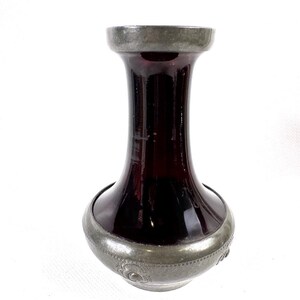 Peut inclure: Un vase vintage avec un corps en verre bordeaux foncé et des accents en métal couleur étain. Le vase a une base bulbeuse, un col fin et un bord évasé. La ferronnerie présente des motifs floraux décoratifs.