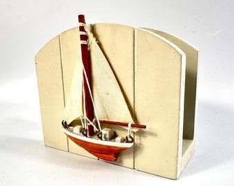 May include: A cream-colored wooden napkin holder featuring a decorative three-dimensional sailboat model attached to the front. The sailboat has a brown hull, a dark red mast, and off-white fabric sails secured with thin white string.