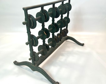 May include: A dark green wooden thread spool rack featuring a rectangular frame with four horizontal bars. Each bar holds four circular spools, totaling sixteen spools arranged in a grid. The structure is supported by two curved feet.