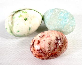 May include: Three smooth, polished stone eggs. One egg is white with green flecks, one is a pale, mottled blue-green, and the third is a variegated pink and deep red stone with a glossy, marbled appearance and rounded, oval shapes.