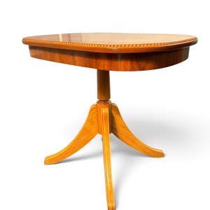 Op de afbeelding: Een lichtbruine houten bijzettafel met een glad, afgerond rechthoekig blad en een gedetailleerde rand met kraalmotief. De tafel rust op een centrale kolom die uitloopt in drie gebogen poten, wat zorgt voor een stabiele basis voor het blad.