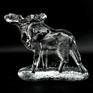 May include: A clear glass moose figurine with detailed antlers and a textured body. The moose stands on a frosted base, with its head raised. The sculpture is set against a black background.