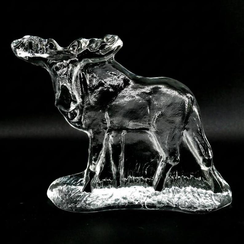 May include: A clear glass moose figurine with detailed antlers and a textured body. The moose stands on a frosted base, with its head raised. The sculpture is set against a black background.