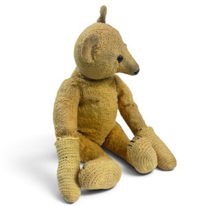 Pode incluir: Um urso de peluche de estilo vintage com pelo castanho-dourado gasto, apresentando um focinho pontiagudo, olhos escuros e uma pequena cauda. O urso tem tecido texturizado tipo croch&eacute; nas patas, com costuras pretas vis&iacute;veis nas solas.