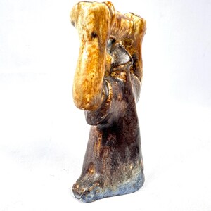 May include: A ceramic sculpture with a textured surface. The sculpture features a figure with raised arms, in shades of brown, tan, and blue. The base has a blue hue. The artwork is set against a white background.