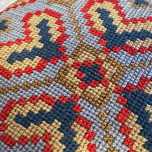 May include: A needlepoint textile featuring a repeating geometric pattern composed of cross-stitched squares. The design utilizes a color palette of vibrant red, deep navy blue, and muted gold threads against a light gray background fabric.