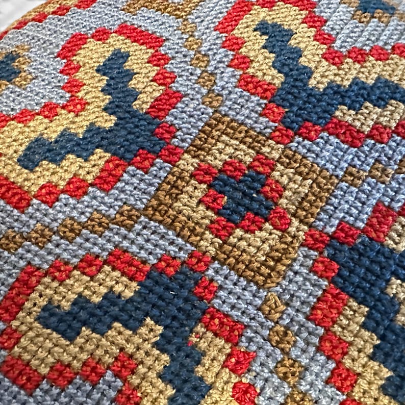 May include: A needlepoint textile featuring a repeating geometric pattern composed of cross-stitched squares. The design utilizes a color palette of vibrant red, deep navy blue, and muted gold threads against a light gray background fabric.
